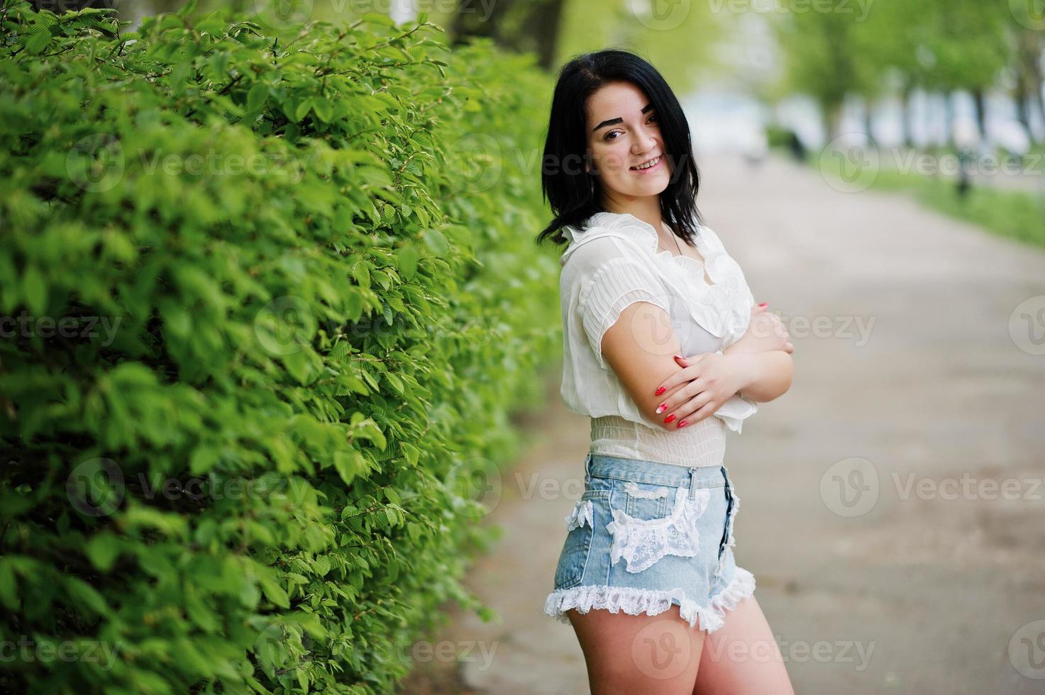 retrato de menina morena sexy em shorts jeans feminino e blusa branca contra arbustos verdes de primavera. foto