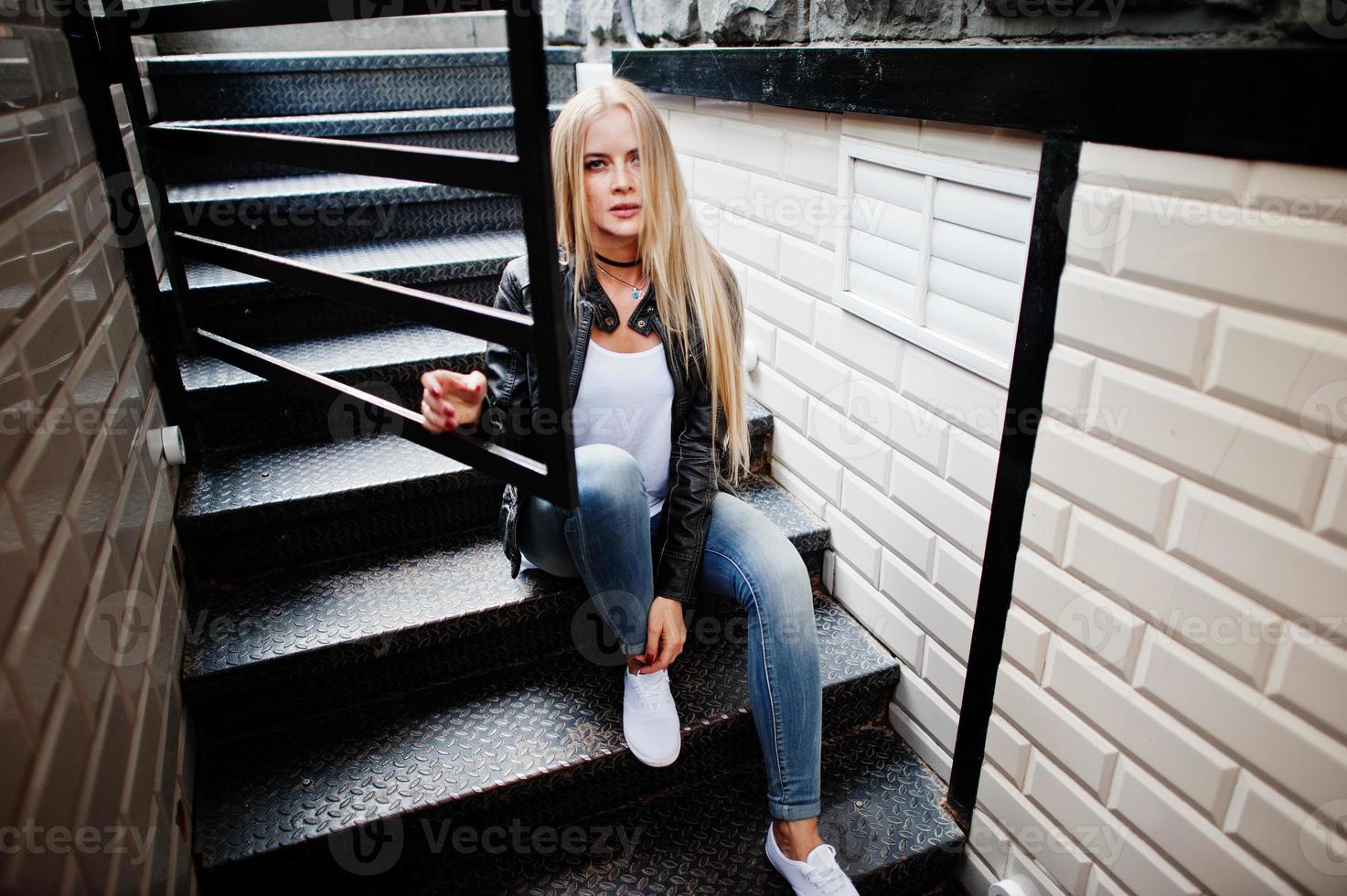 mulher loira elegante usa jeans, gargantilha e jaqueta de couro na rua perto de escadas. retrato de modelo urbano de moda. foto