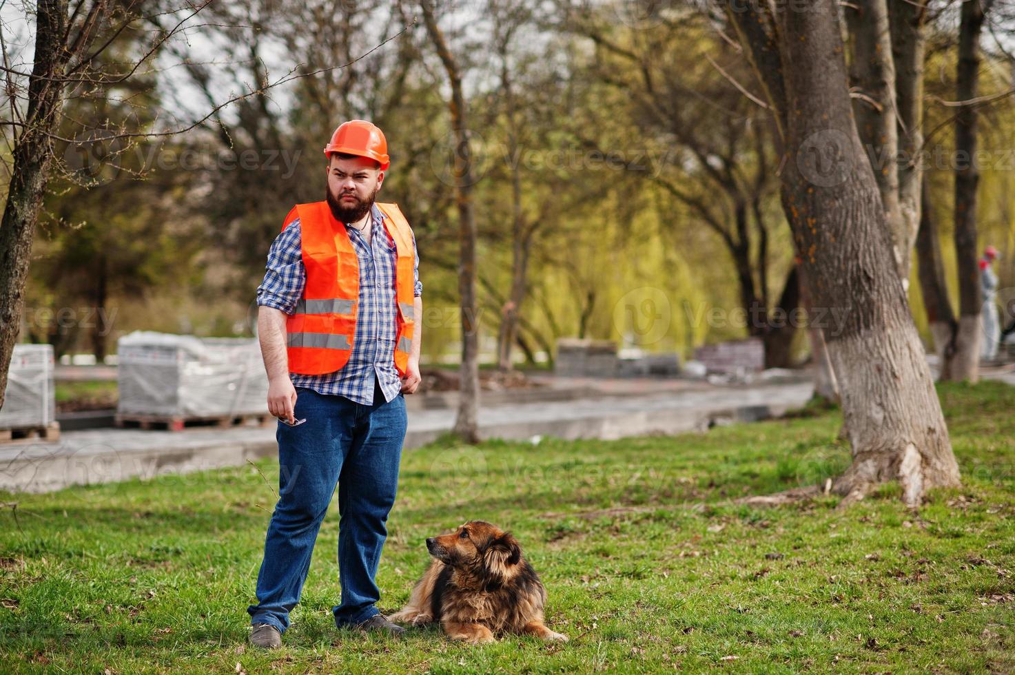 retrato de trabalhador de barba brutal homem terno trabalhador da construção civil no capacete laranja de segurança com cão de guarda. foto