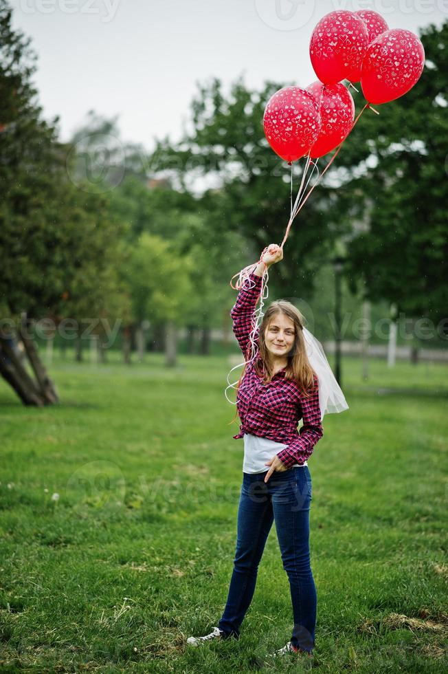 retrato de menina morena na camisa quadriculada, jeans e véu com muitos balões vermelhos na festa de despedida. foto