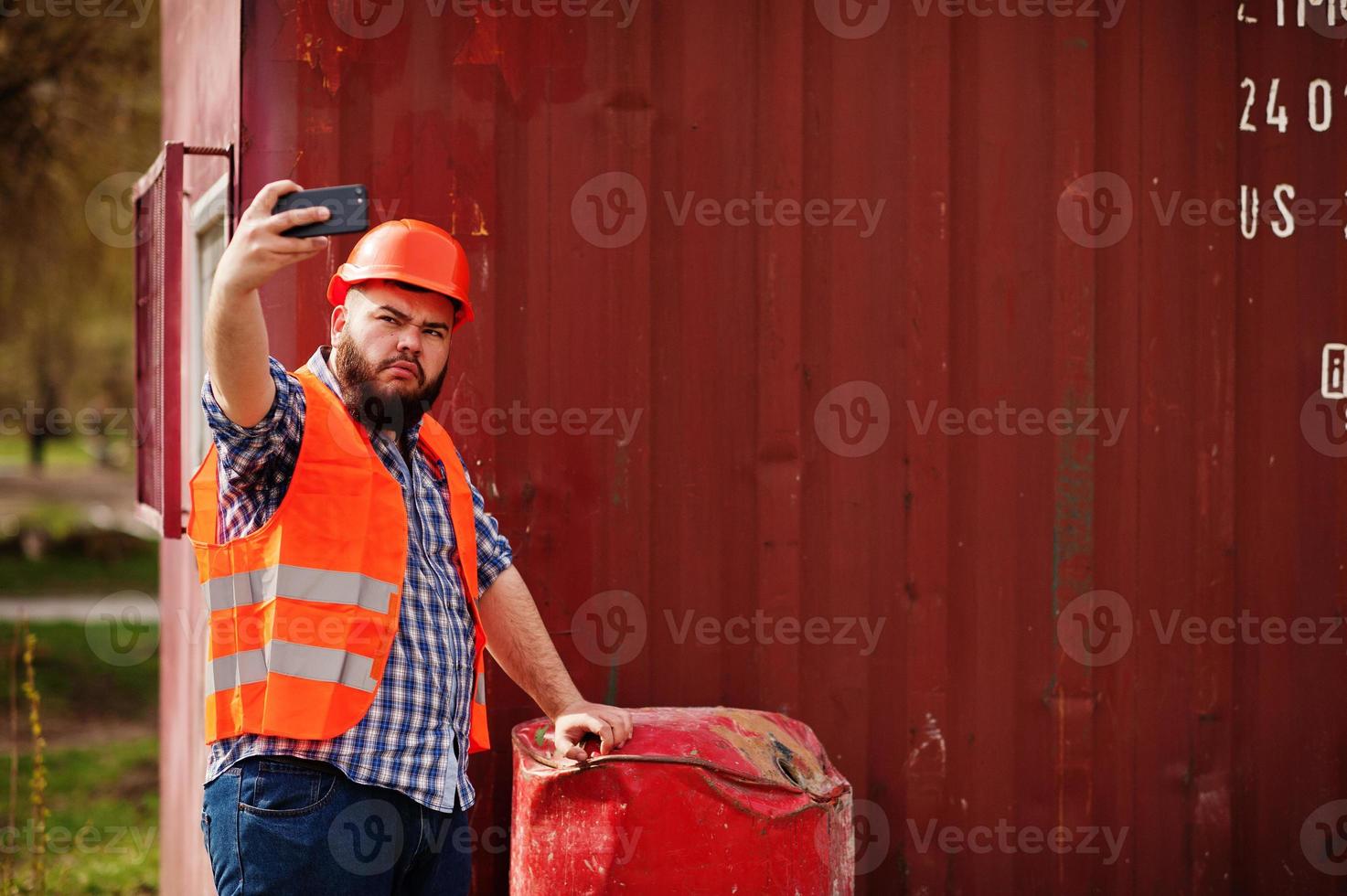 trabalhador de barba homem terno trabalhador da construção civil no capacete laranja de segurança perto do barril vermelho faz selfie no telefone. foto