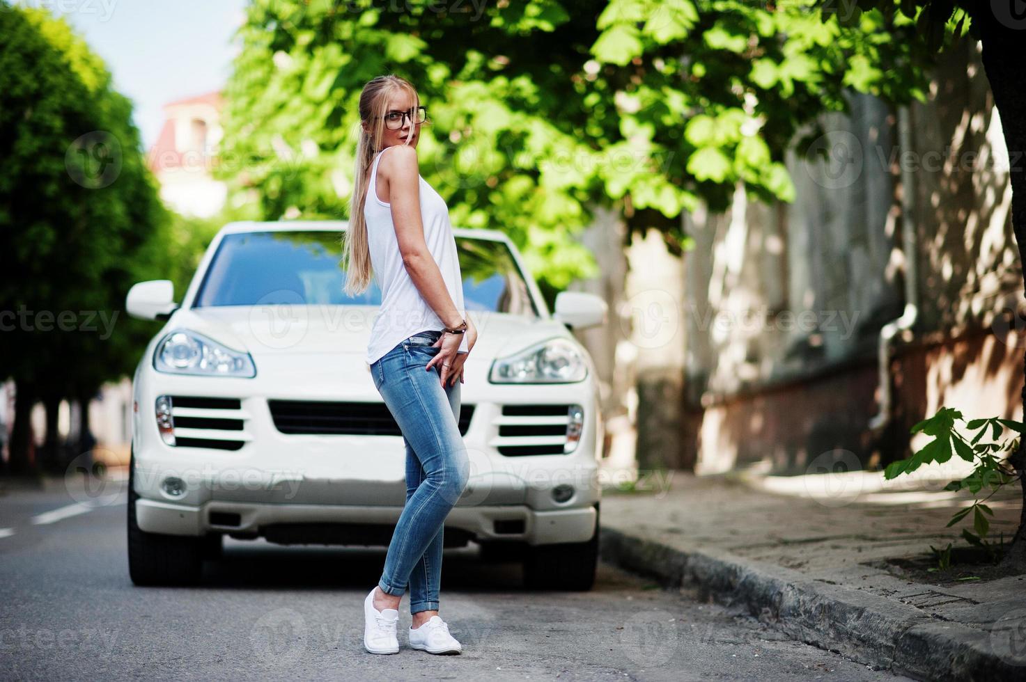 mulher loira elegante usa jeans, óculos, gargantilha e camisa branca contra carro de luxo. retrato de modelo urbano de moda. foto