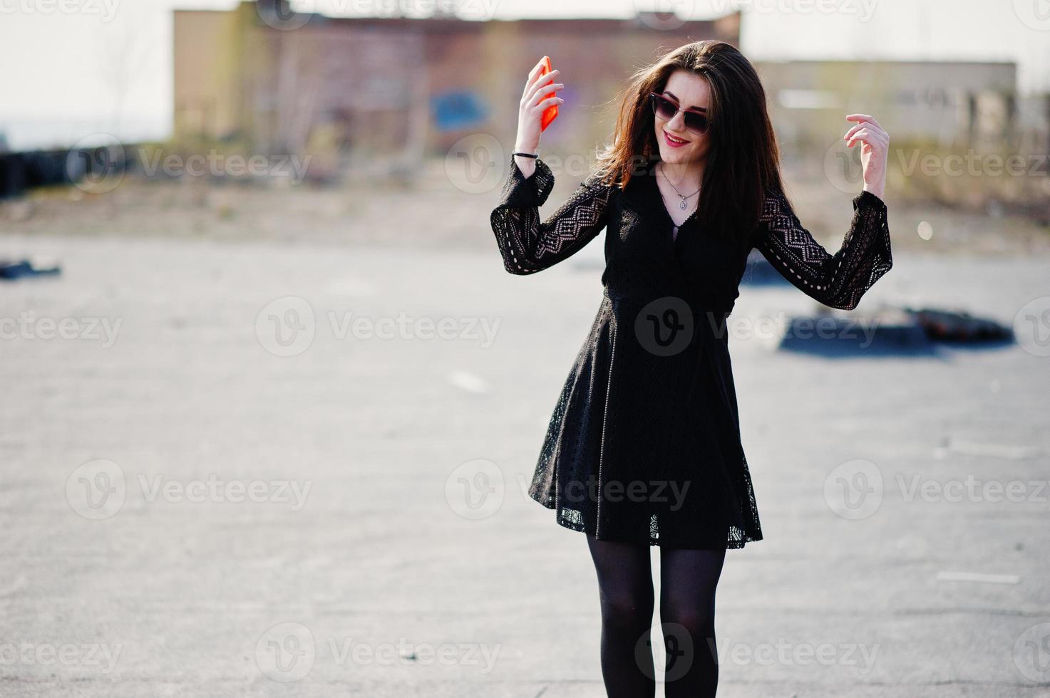 menina morena retrato com lábios vermelhos e celular laranja nas mãos, vestindo um vestido preto, óculos de sol posou no telhado. modelo de moda de rua. foto