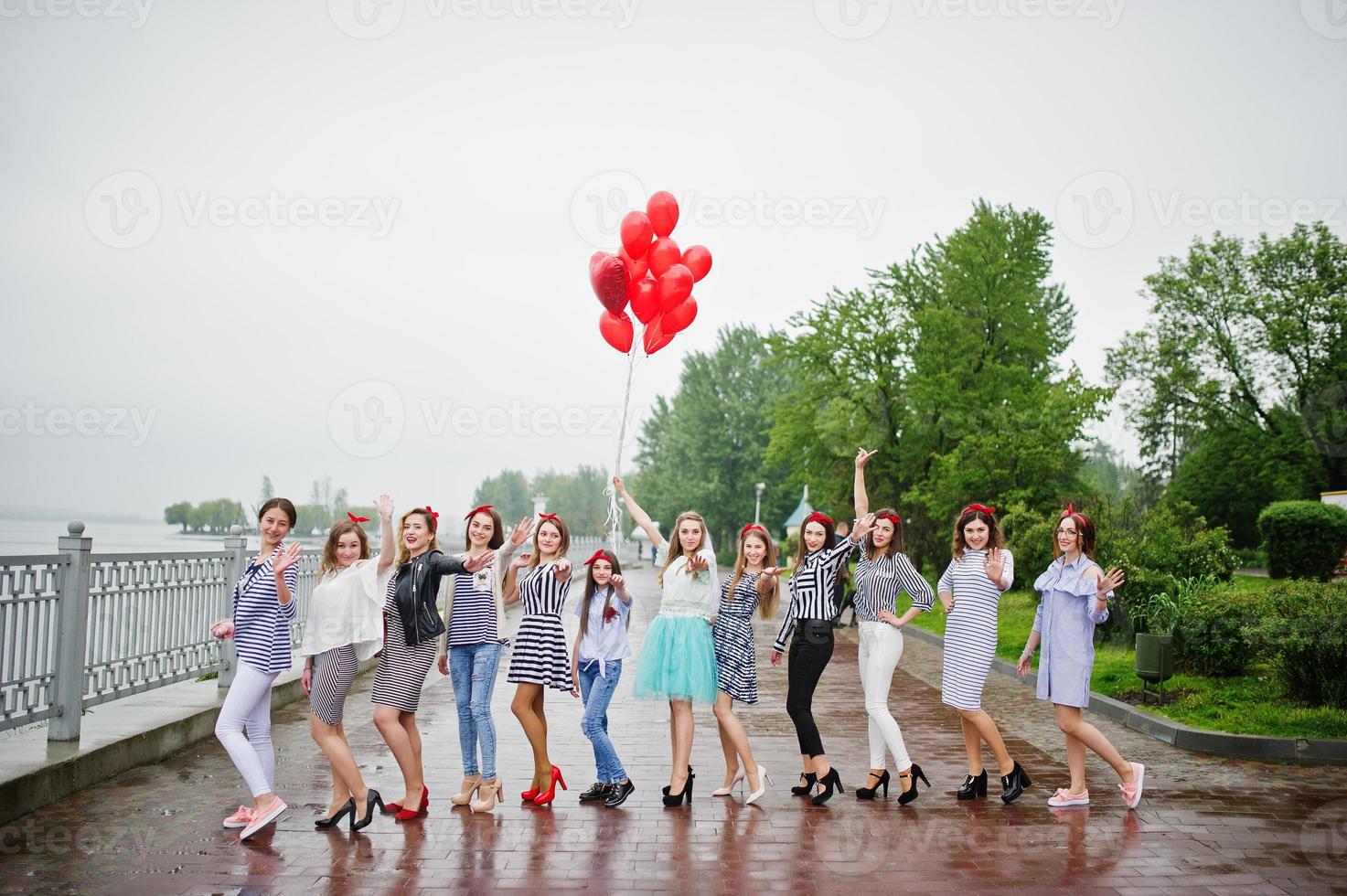 onze damas de honra incrivelmente bonitas com noiva deslumbrante posando com balões vermelhos em forma de coração na calçada contra o lago ao fundo. foto