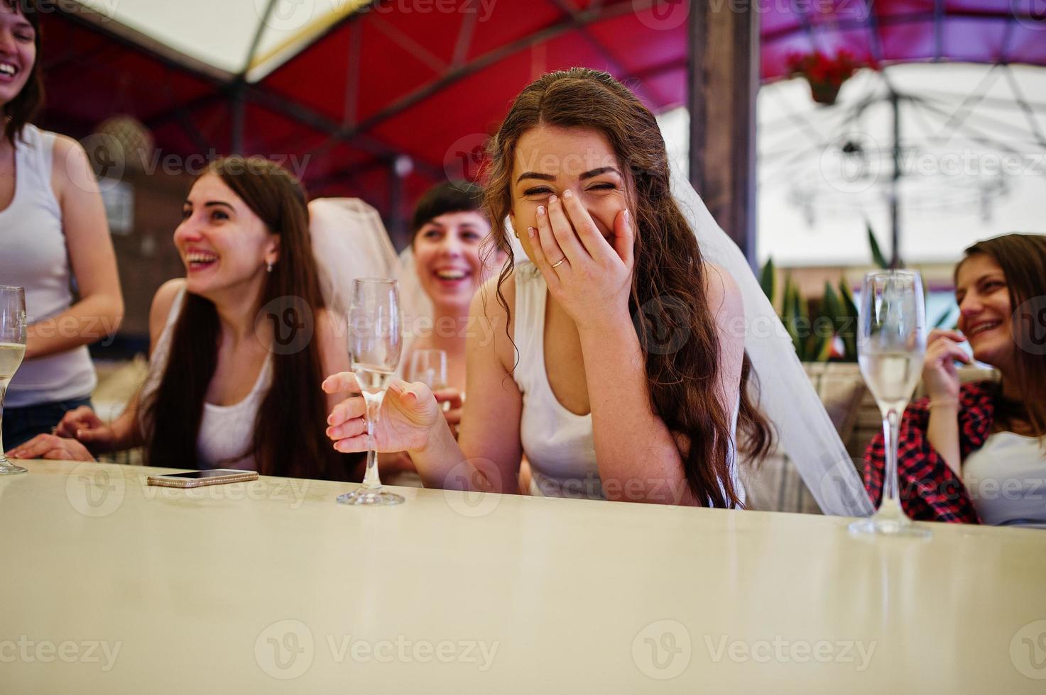 grupo de meninas alegres em camisas brancas, sentado à mesa e beber champanhe na festa de despedida. foto