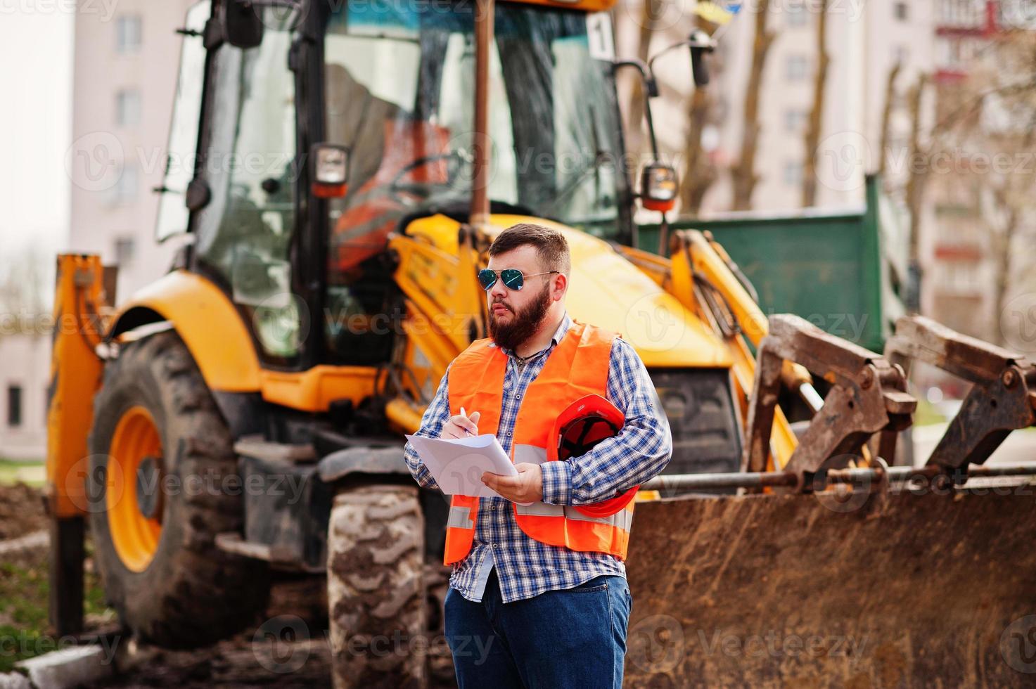 homem trabalhador barba brutal terno trabalhador da construção civil no capacete laranja de segurança, óculos de sol contra traktor com papel plano nas mãos. foto