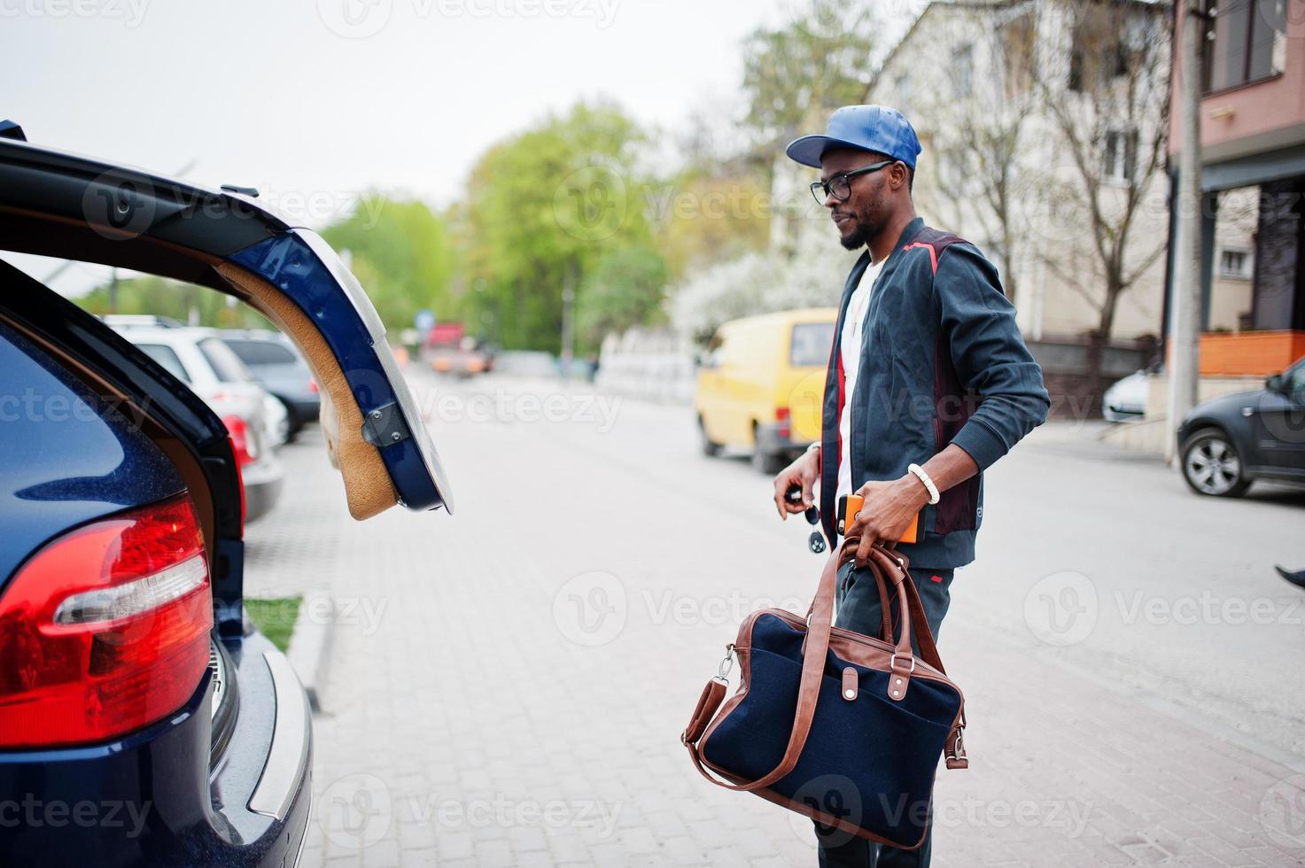 retrato de homem afro-americano elegante em roupas esportivas, boné e óculos andando com bolsa e porta-malas de carro aberto. homens negros modelam moda de rua. foto