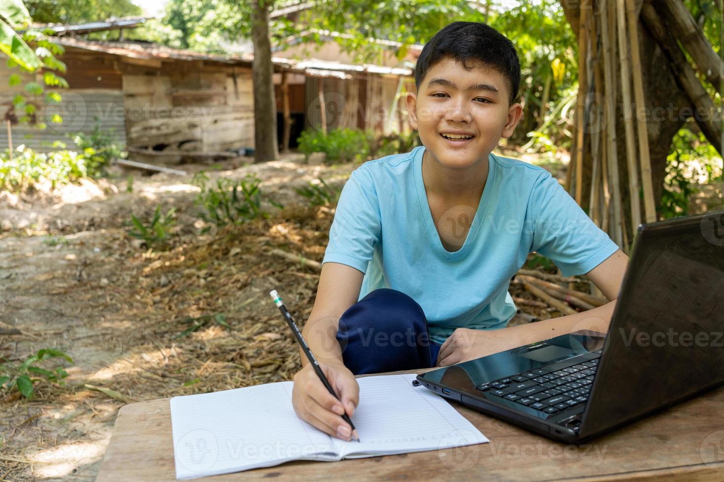 menino olhando para a câmera durante as aulas online alegremente com o computador portátil em casa no campo foto