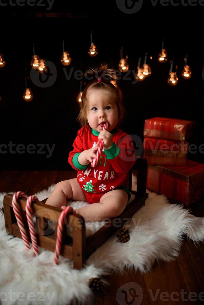 Menina de 7 meses em uma fantasia de natal vermelha em um fundo de guirlandas retrô senta-se em uma pele foto