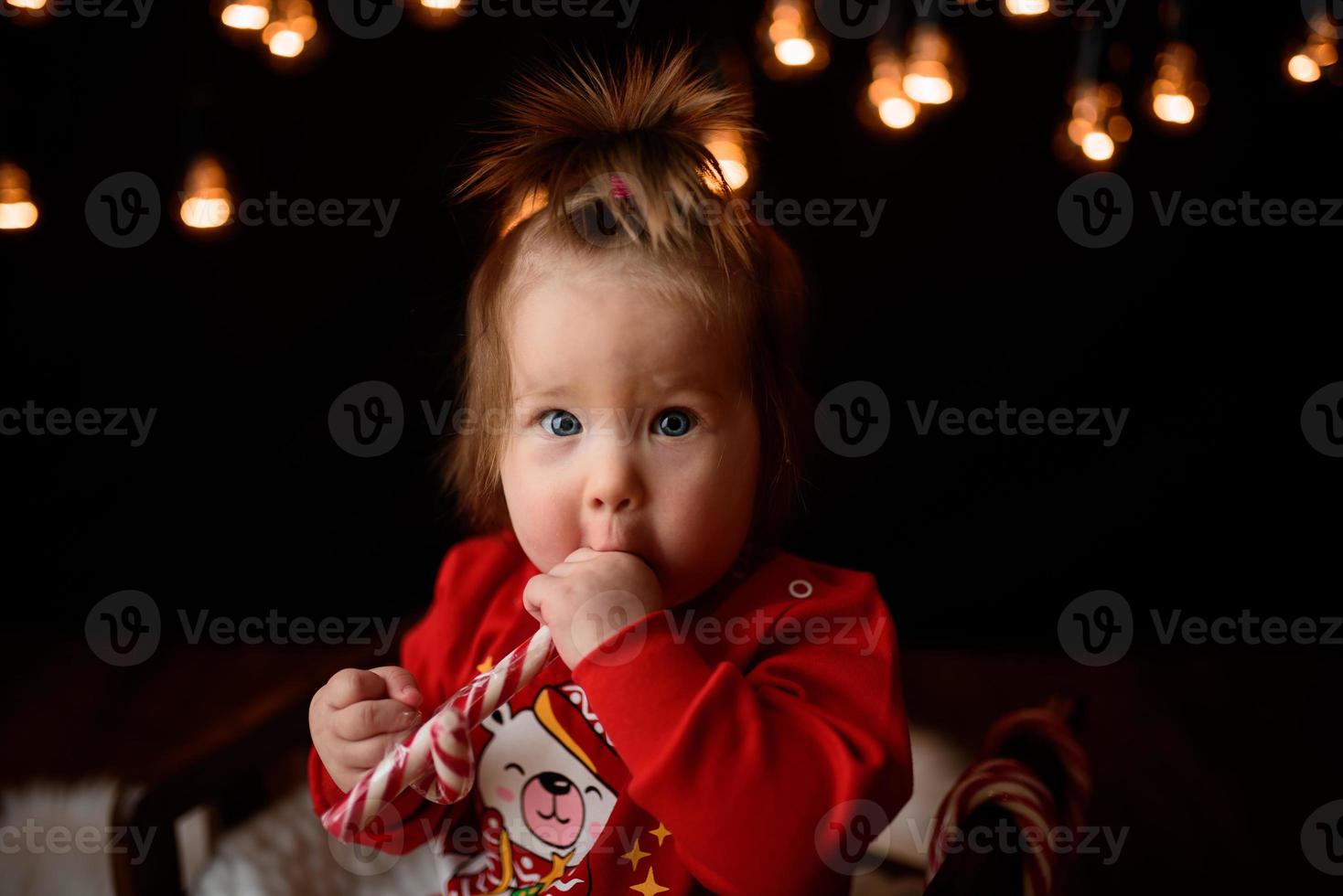 Menina de 7 meses em uma fantasia de natal vermelha em um fundo de guirlandas retrô senta-se em uma pele foto