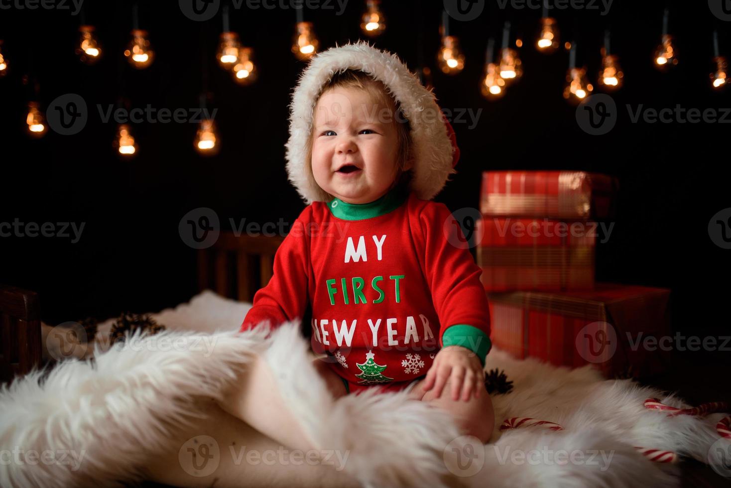 Menina de 7 meses em uma fantasia de natal vermelha em um fundo de guirlandas retrô senta-se em uma pele foto