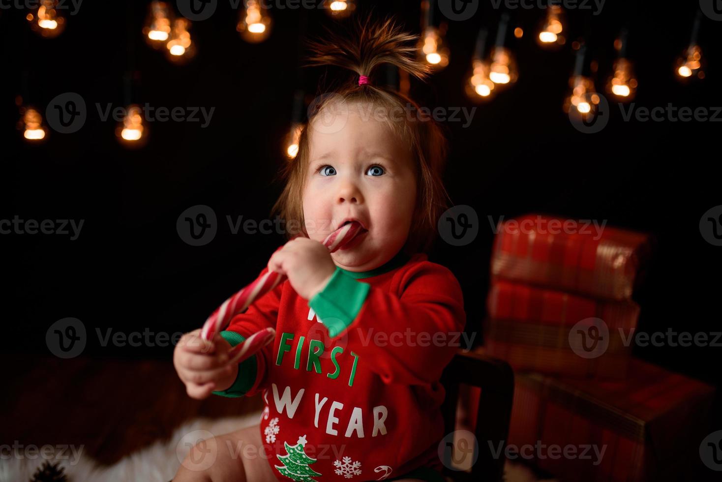 Menina de 7 meses em uma fantasia de natal vermelha em um fundo de guirlandas retrô senta-se em uma pele foto