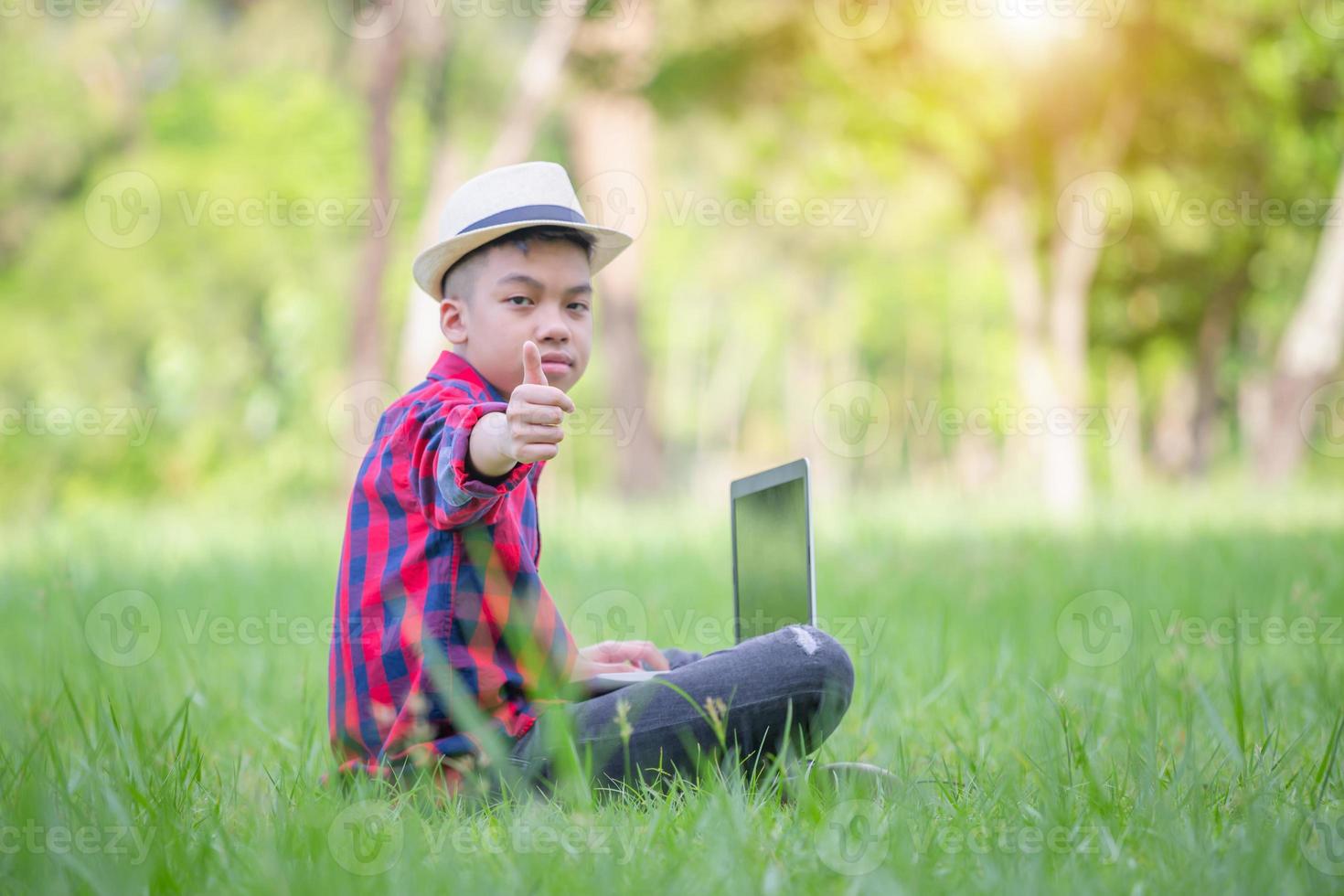 menino dando polegares para cima e sentado na grama estudando com laptop no parque, conceito de aprendizagem ao ar livre foto