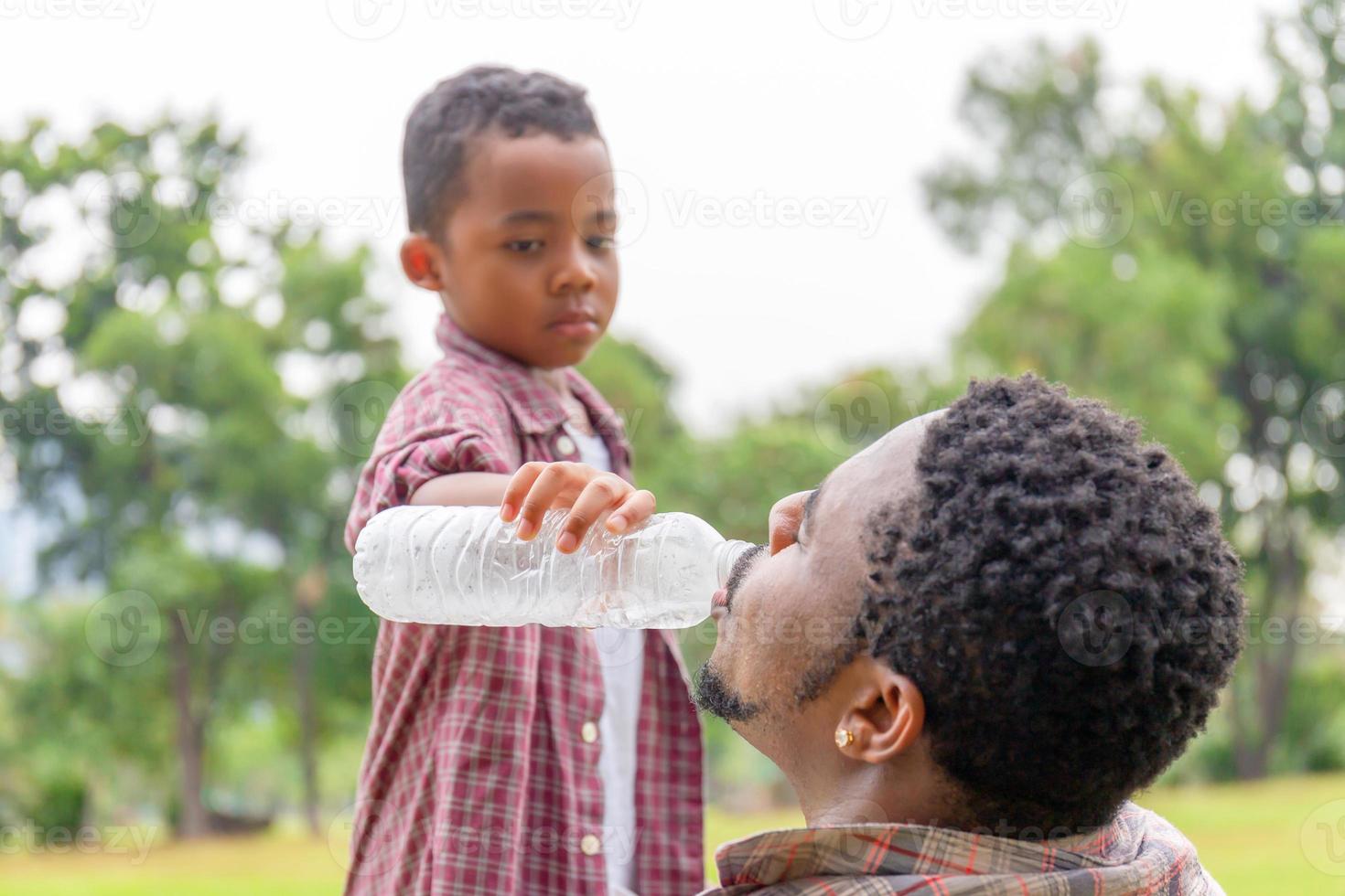 filho dando água para seu pai, pai afro-americano e filho ao ar livre, família de felicidade em conceitos de parque foto