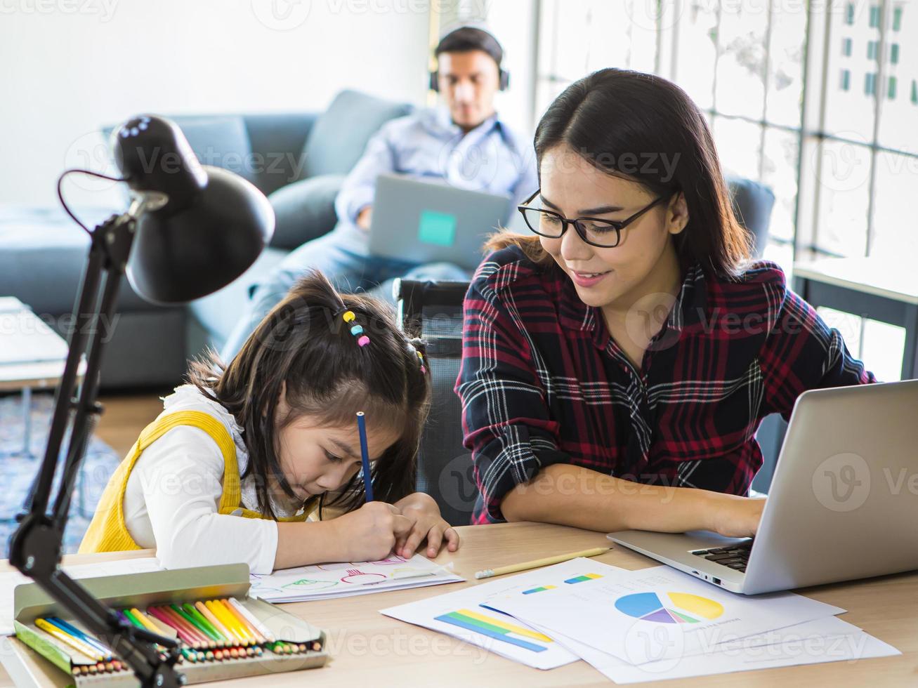 família étnica muti, pai e mãe e filha ficam juntos na sala de estar. mãe trabalha em casa com o computador portátil enquanto ensina a garota a fazer a lição de casa. novo conceito normal foto