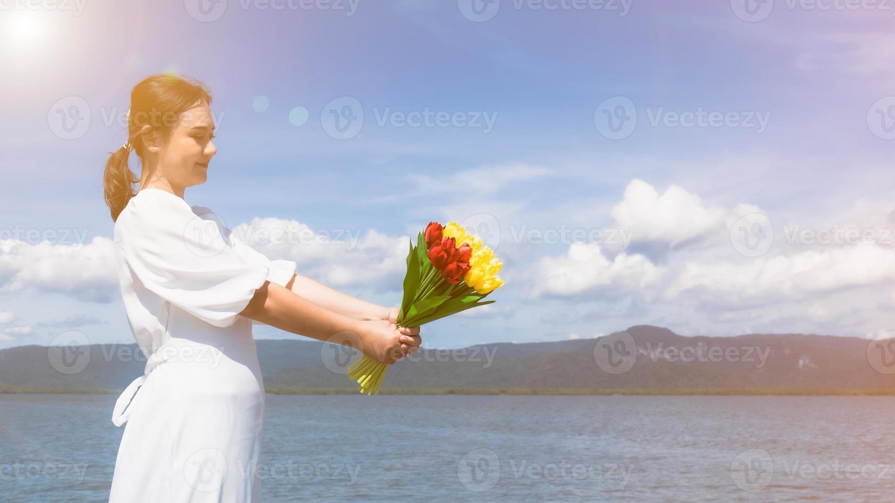 uma jovem asiática de vestido branco segura um buquê de flores amarelas e vermelhas no porto marítimo. à luz do sol, o céu está claro, o vento sopra. foto