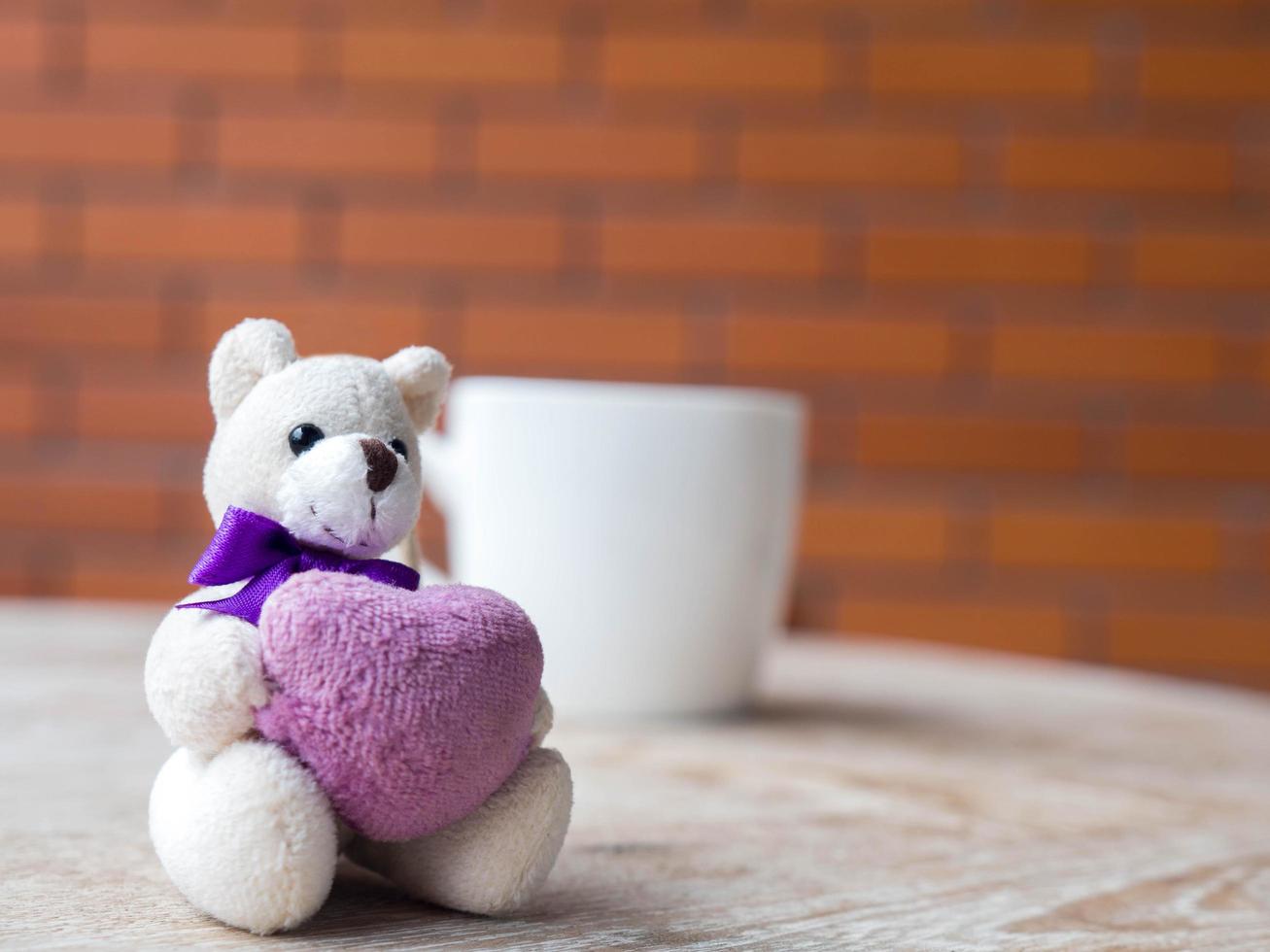 ursinho segurando um coração roxo. viver em uma xícara de café branco. colocado sobre uma mesa de madeira. o pano de fundo é um bloco de tijolos marrom. foto