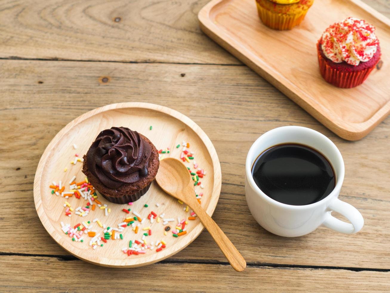 cupcake de chocolate colocado em uma placa de madeira esférica. ao lado do cupcake tem despertador vintage e caneca de café branco. no fundo tem cupcake amarelo e cupcakes vermelhos. foto