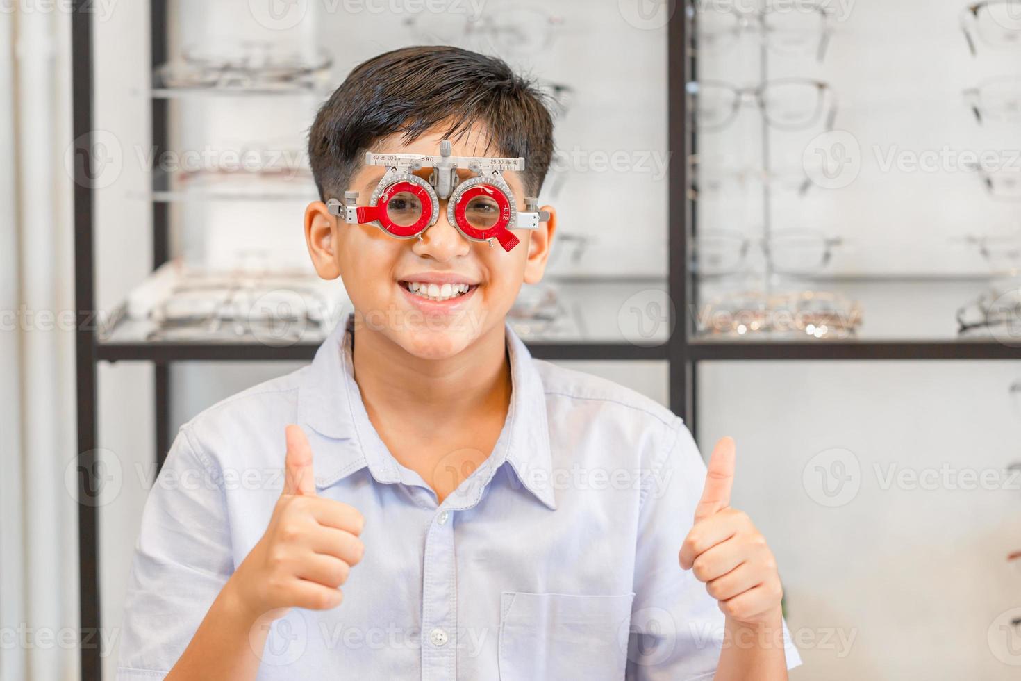 sorridente menino indiano-tailandês escolhendo óculos na loja de óptica, retrato de criança usando óculos de teste de olho na loja de óptica mostrando os polegares foto