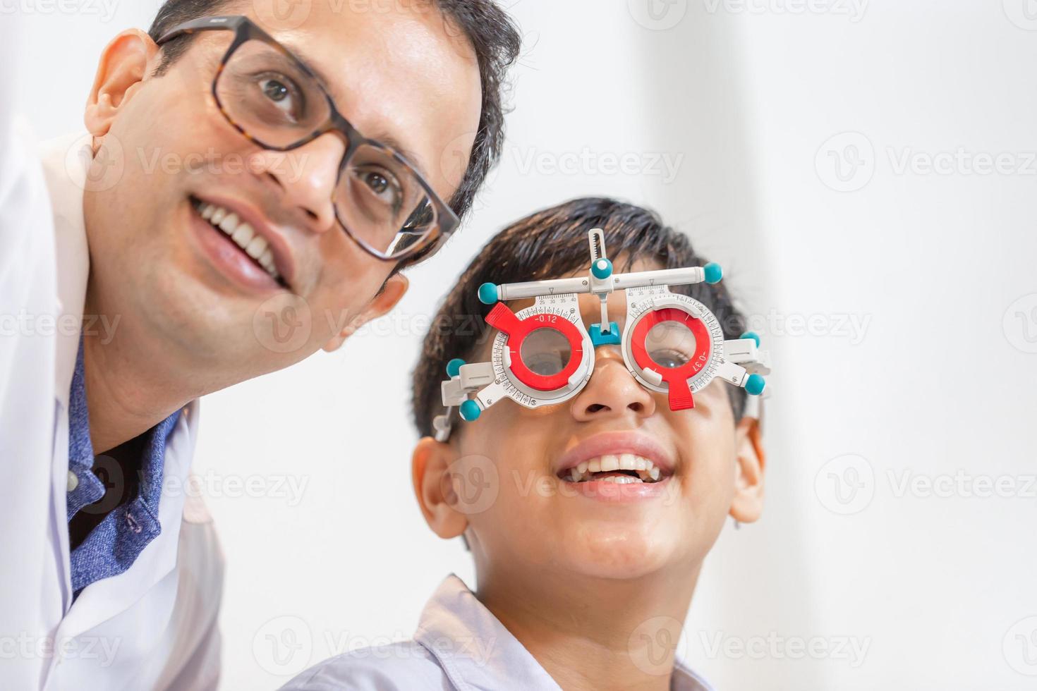 menino indiano-tailandês sorridente escolhendo óculos na loja de óptica, menino fazendo exame de verificação de olho com optometrista usando armação de teste na loja de óptica foto
