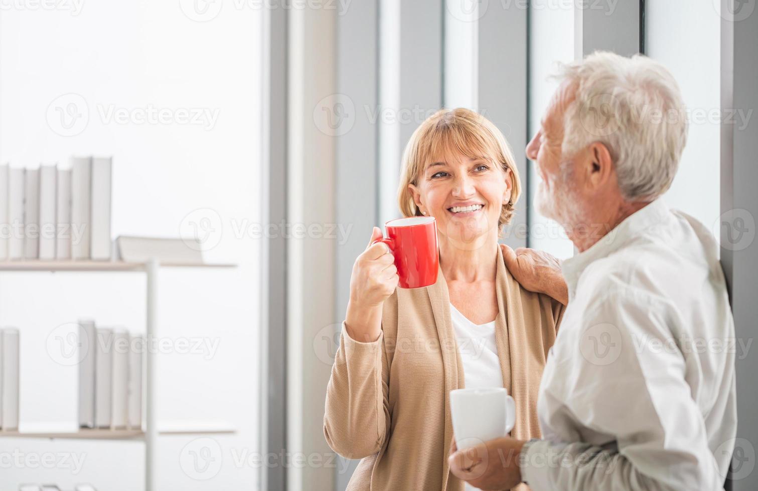 casal sênior dentro de nova casa durante o intervalo para o café, casal de idosos conversando em pé perto da janela com xícaras de café foto