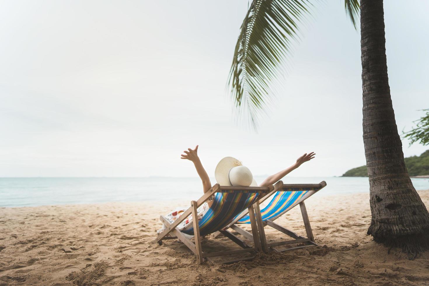 jovem mulher da ásia relaxando na cadeira de praia braço para cima a mão com chapéu flexível foto