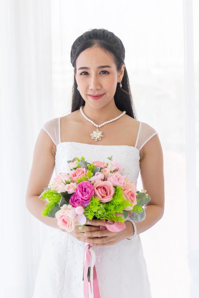 uma noiva asiática vestida com um vestido de noiva branco fica sorrindo brilhantemente em uma mão segurando um lindo buquê de flores. foto