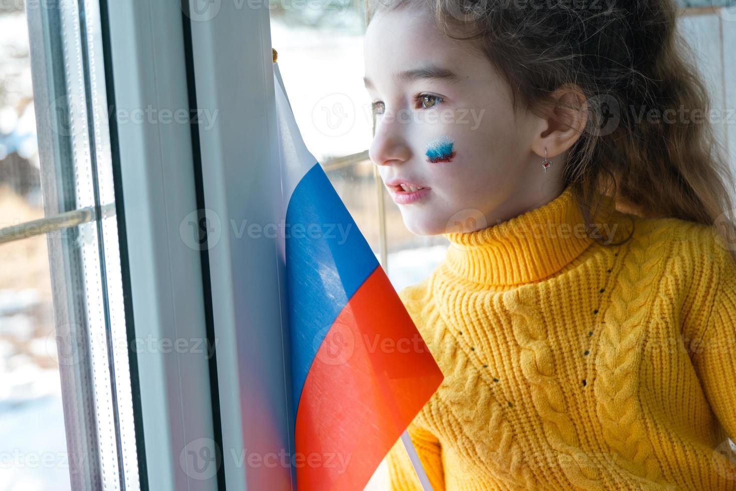 uma criança triste na janela com a bandeira da rússia, se preocupa com lágrimas nos olhos. conflito entre rússia e ucrânia, medo foto