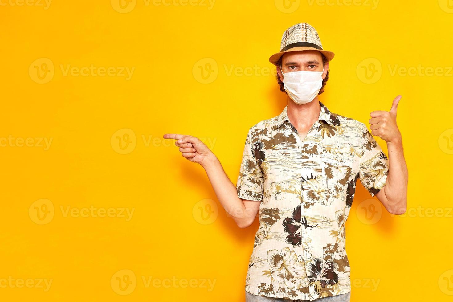 turista de férias masculino com máscara médica aponta o dedo para uma área vazia e fica com o polegar levantado isolado em fundo amarelo, vestido de camisa, chapéu. descanso de conceito, férias, viagens foto