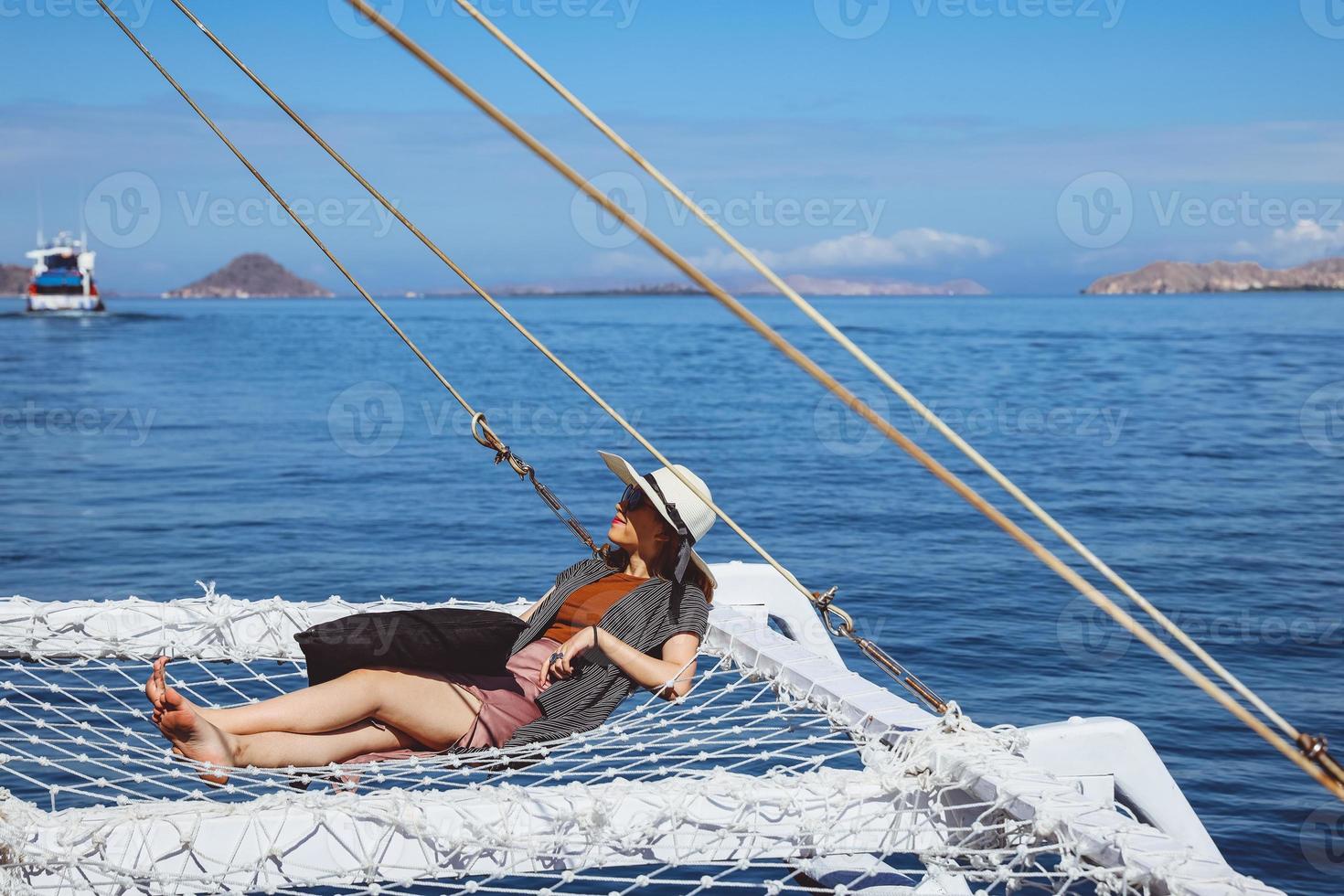 mulher de chapéu de verão e óculos de sol relaxando na rede de catamarã, apreciando a vista para o mar foto