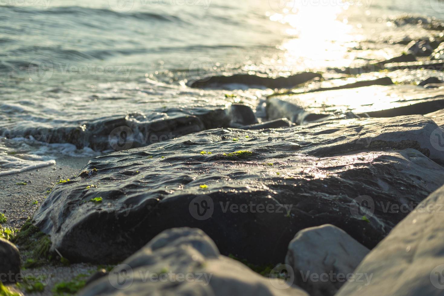 rochas brilhantes e ondas quebrando à beira-mar com brilho do sol e algas marinhas foto