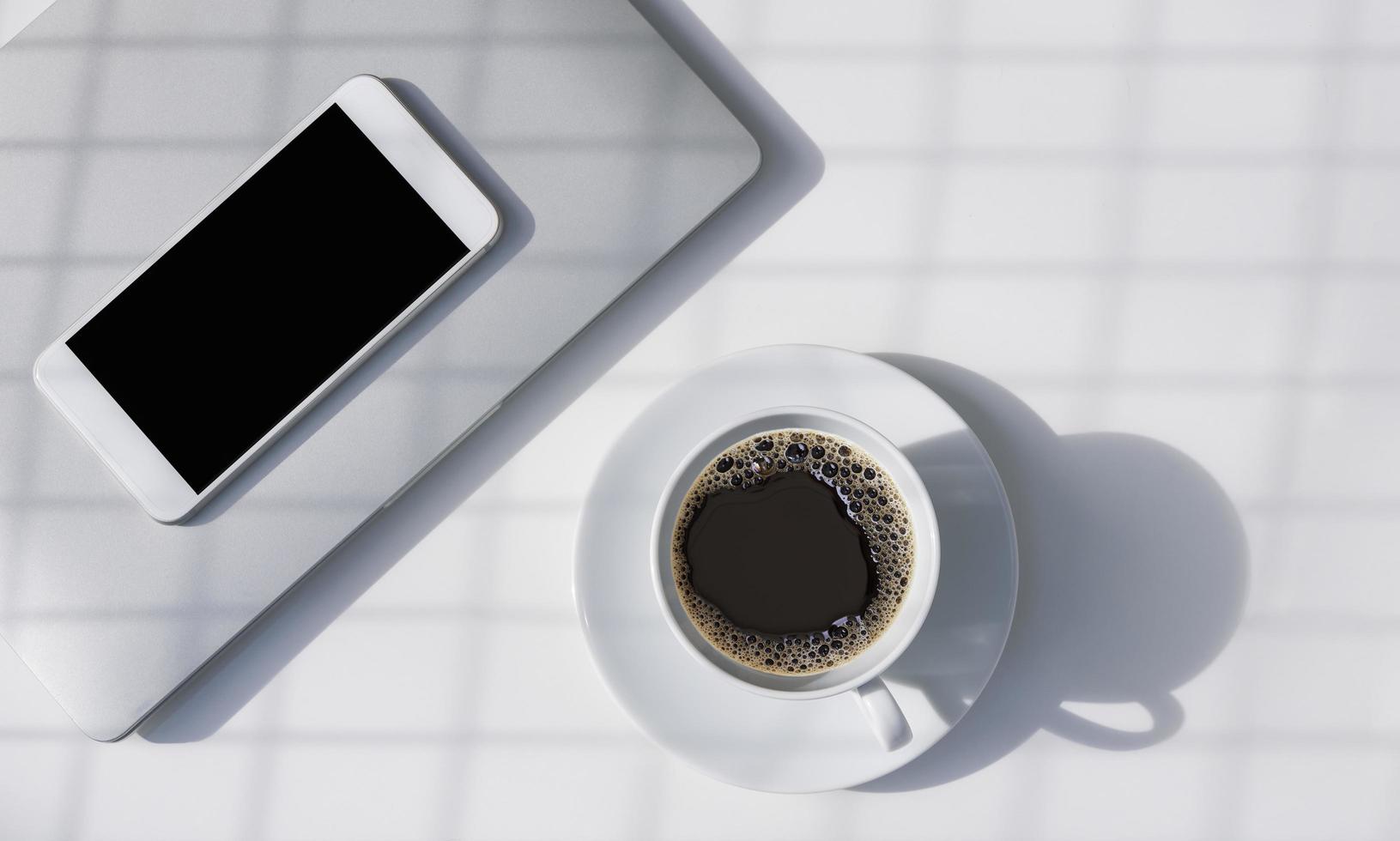 padrão de sombra de luz solar e grade na superfície do café preto em xícara de cerâmica branca com telefone inteligente em branco e laptop cinza na mesa branca foto