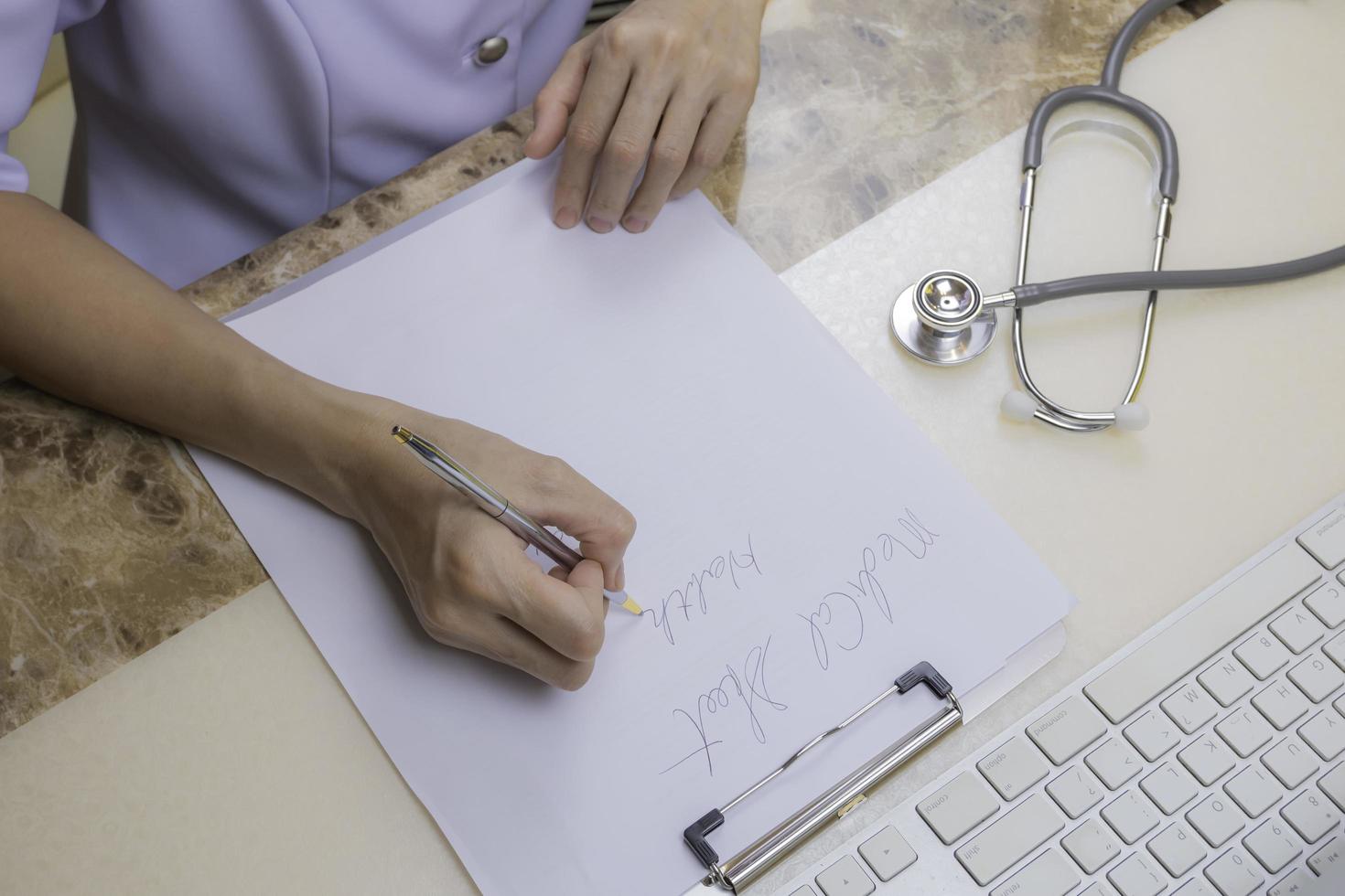 vista de alto ângulo da médica ou enfermeira asiática está escrevendo prescrição com estetoscópio e parte do teclado do computador em cima da mesa na sala de escritório do hospital foto