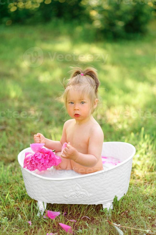 menina criança feliz toma um banho de leite com pétalas. menina em um banho de leite sobre um fundo verde. buquês de peônias rosa. banho do bebê. higiene e cuidados com as crianças. foto