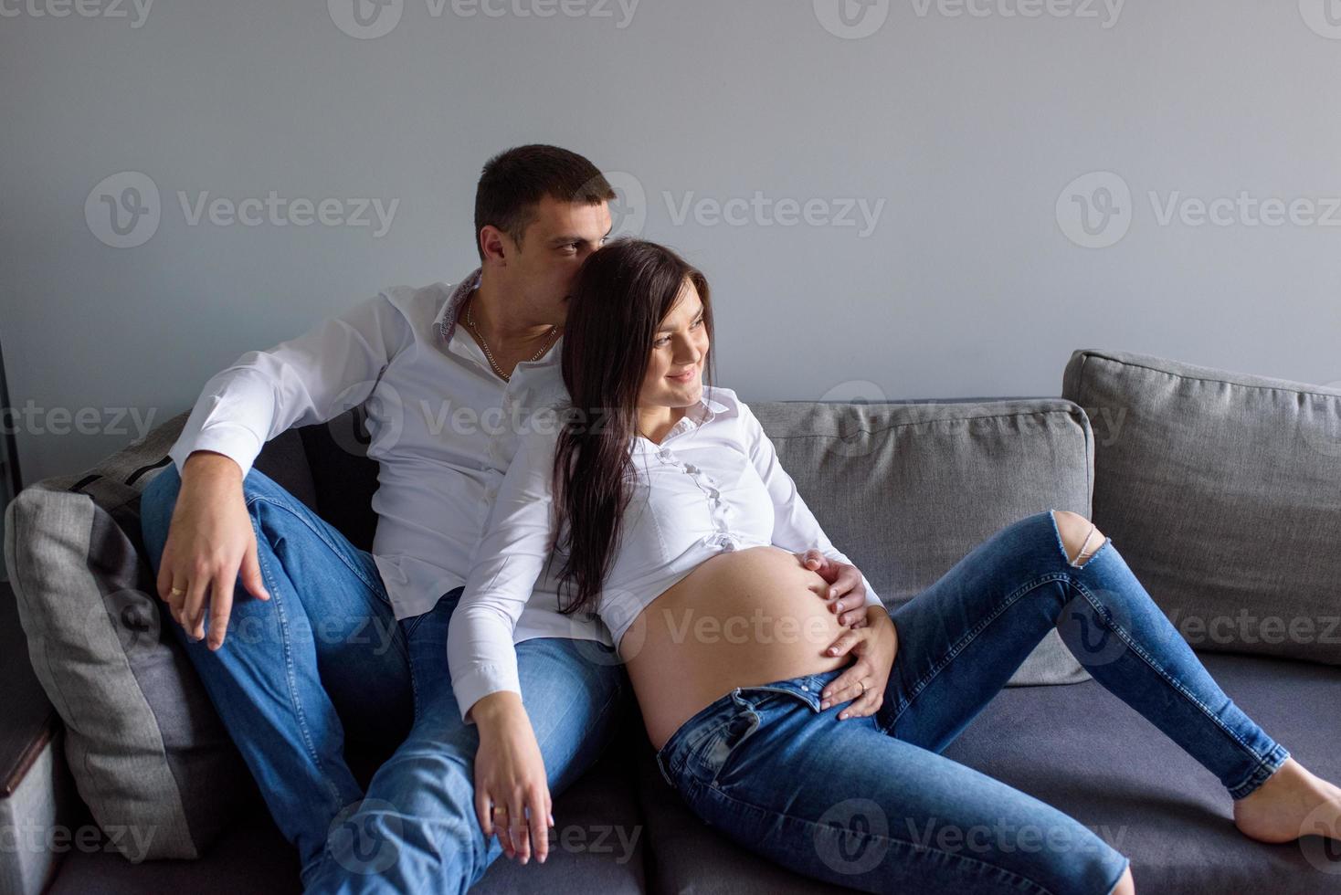 um homem e uma mulher grávida estão sentados abraçados no sofá. foto
