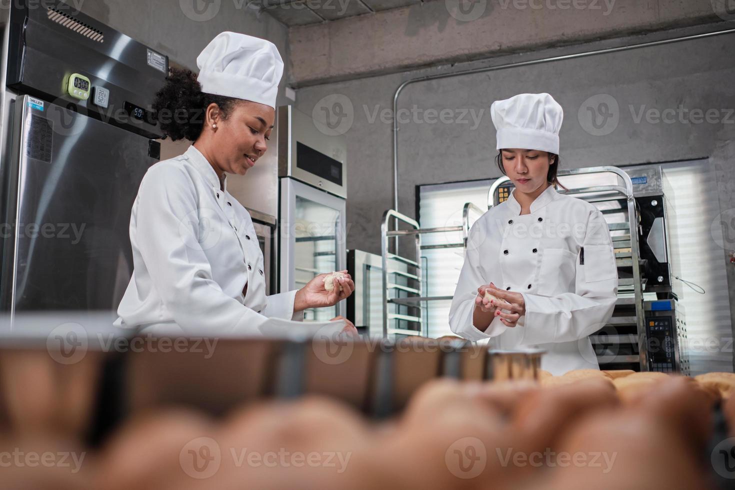 duas chefs profissionais em uniformes e aventais de cozinheiro branco amassam massa de pastelaria e ovos, preparam pão, biscoitos e alimentos frescos de padaria, assando no forno em uma cozinha de aço inoxidável de um restaurante. foto