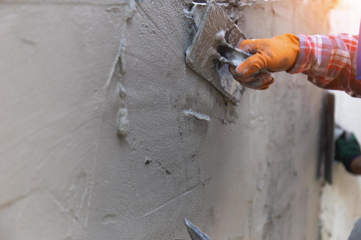 as mãos de um trabalhador que está rebocando de perto estão usando luvas de borracha laranja para evitar que o cimento morda suas mãos, construindo as paredes da casa e tendo belas luzes laranja. foto