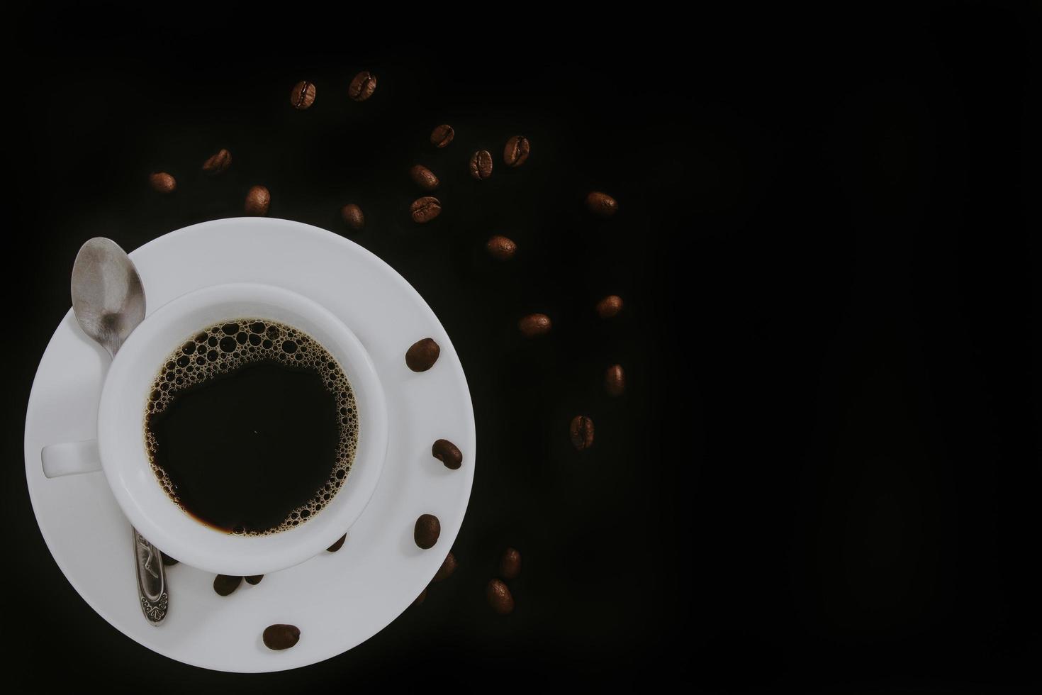 café preto na xícara de café branco e pires com café em grãos, colher de aço inoxidável em fundo preto. foto
