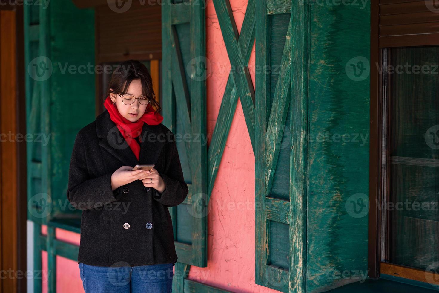uma garota com um telefone nas mãos fica na rua perto de uma casa rosa com persianas verdes foto