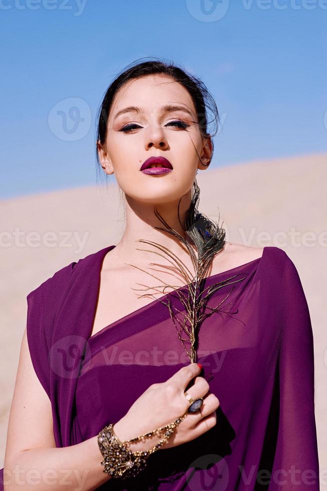 incrível linda mulher morena com a pena de pavão em tecido roxo no deserto. oriental, indiano, moda, conceito de estilo foto