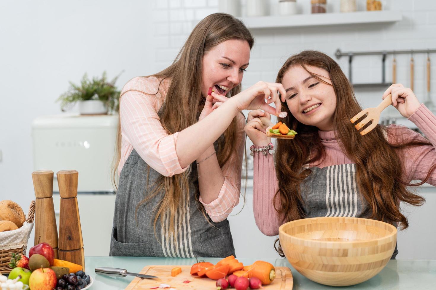 feliz mãe e filha gostam de cozinhar salada caseira na cozinha. garota sorridente preparando legumes. atividade infantil com os pais em casa foto