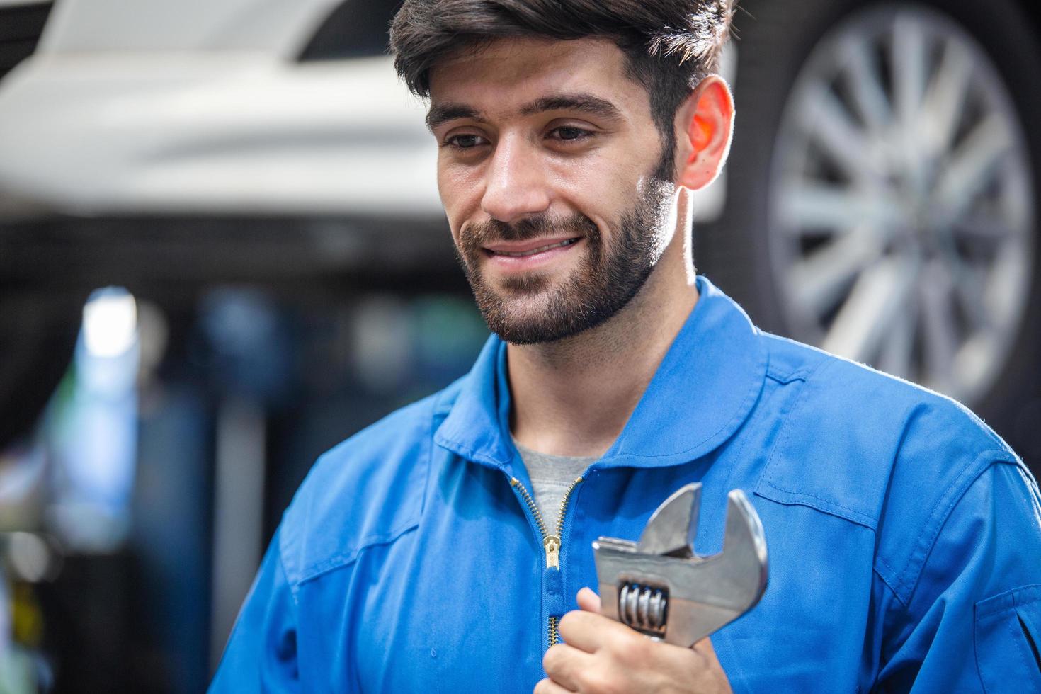 sorridente mecânico masculino caucasiano segurando a chave na garagem de automóveis. carreira de reparo e manutenção. após o serviço, verificando a parte quebrada do dano do carro foto