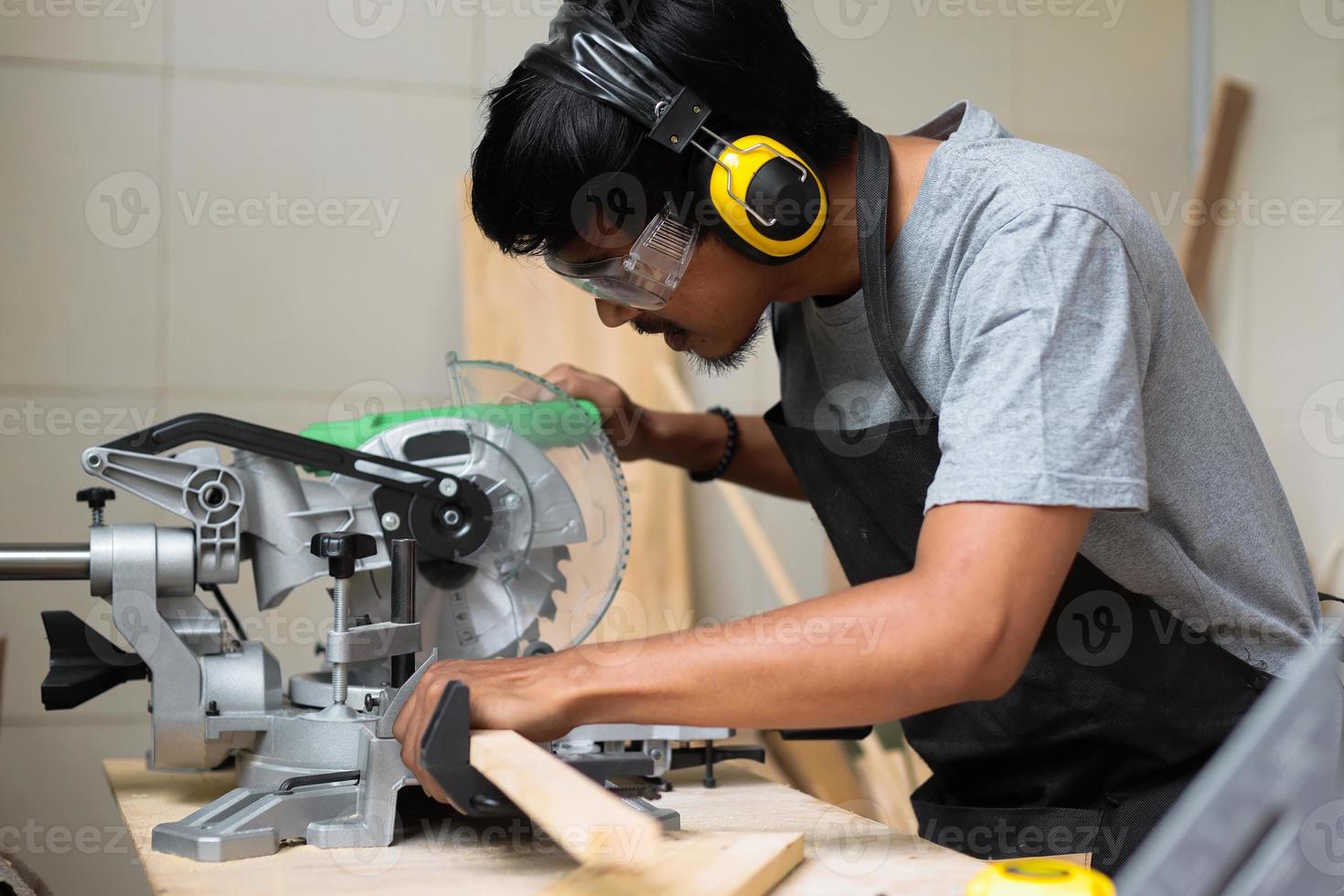 um jovem carpinteiro masculino trabalhando em sua mesa de oficina usando serra circular e usando equipamentos de segurança foto