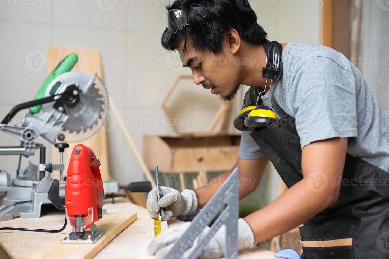 um jovem carpinteiro masculino medindo a madeira em sua mesa de oficina usando equipamentos de segurança foto