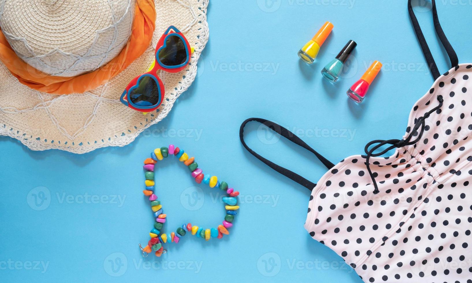 composição de verão. óculos de sol, chapéu, maiô de bolinhas, esmalte e um coração feito de miçangas em um fundo azul com espaço de cópia. conceito de férias., postura plana. foto