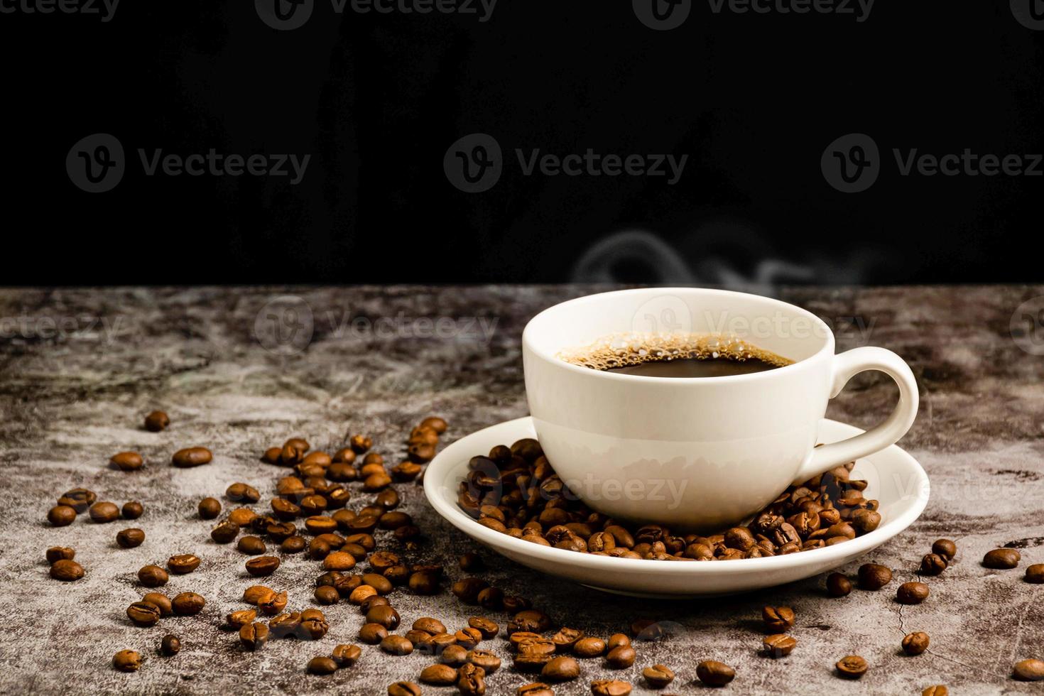 close-up de café quente em uma xícara branca é colocado na mesa de chão de cimento, muitos grãos de café torrados estão no pires da xícara de café e, ao redor, fumaça e aroma flutuam da xícara. fundo preto e desfocado foto