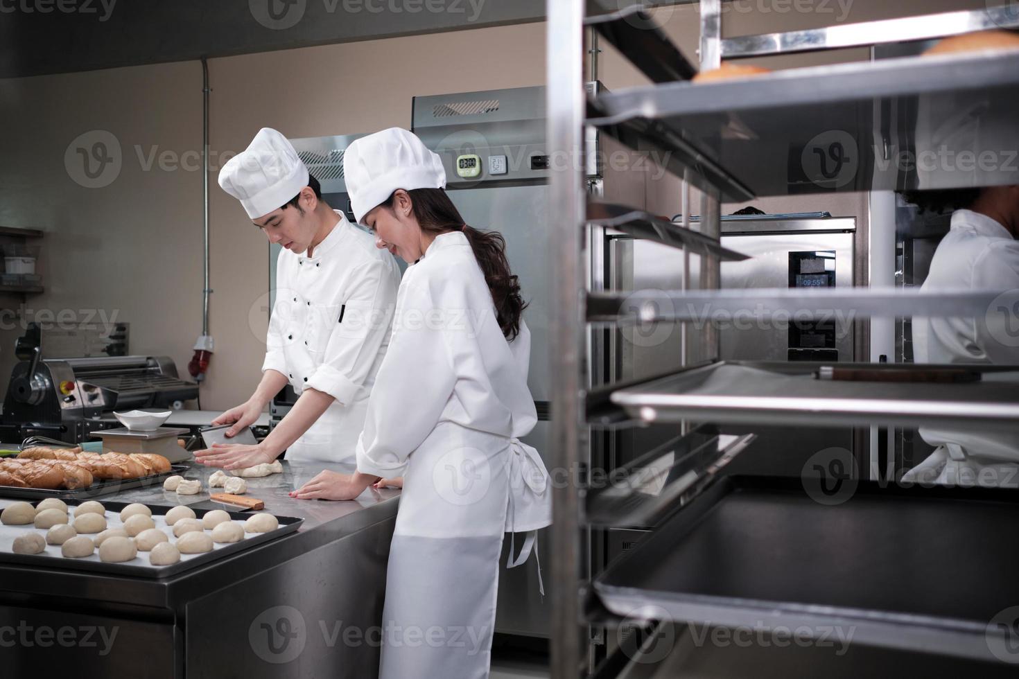 dois chefs asiáticos profissionais em uniformes e aventais de cozinheiro branco amassam massa e ovos, preparam pão, biscoitos e alimentos frescos de padaria, assando em um forno em uma cozinha de aço inoxidável de restaurante. foto