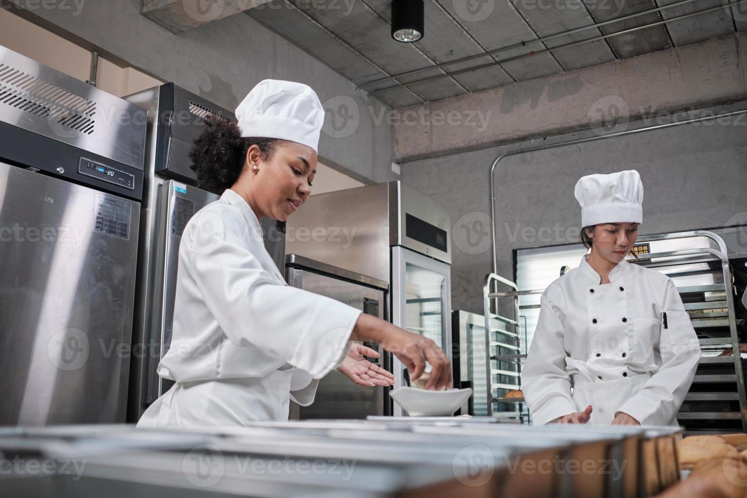 duas chefs profissionais em uniformes e aventais de cozinheiro branco amassam massa de pastelaria e ovos, preparam pão, biscoitos e alimentos frescos de padaria, assando no forno em uma cozinha de aço inoxidável de um restaurante. foto