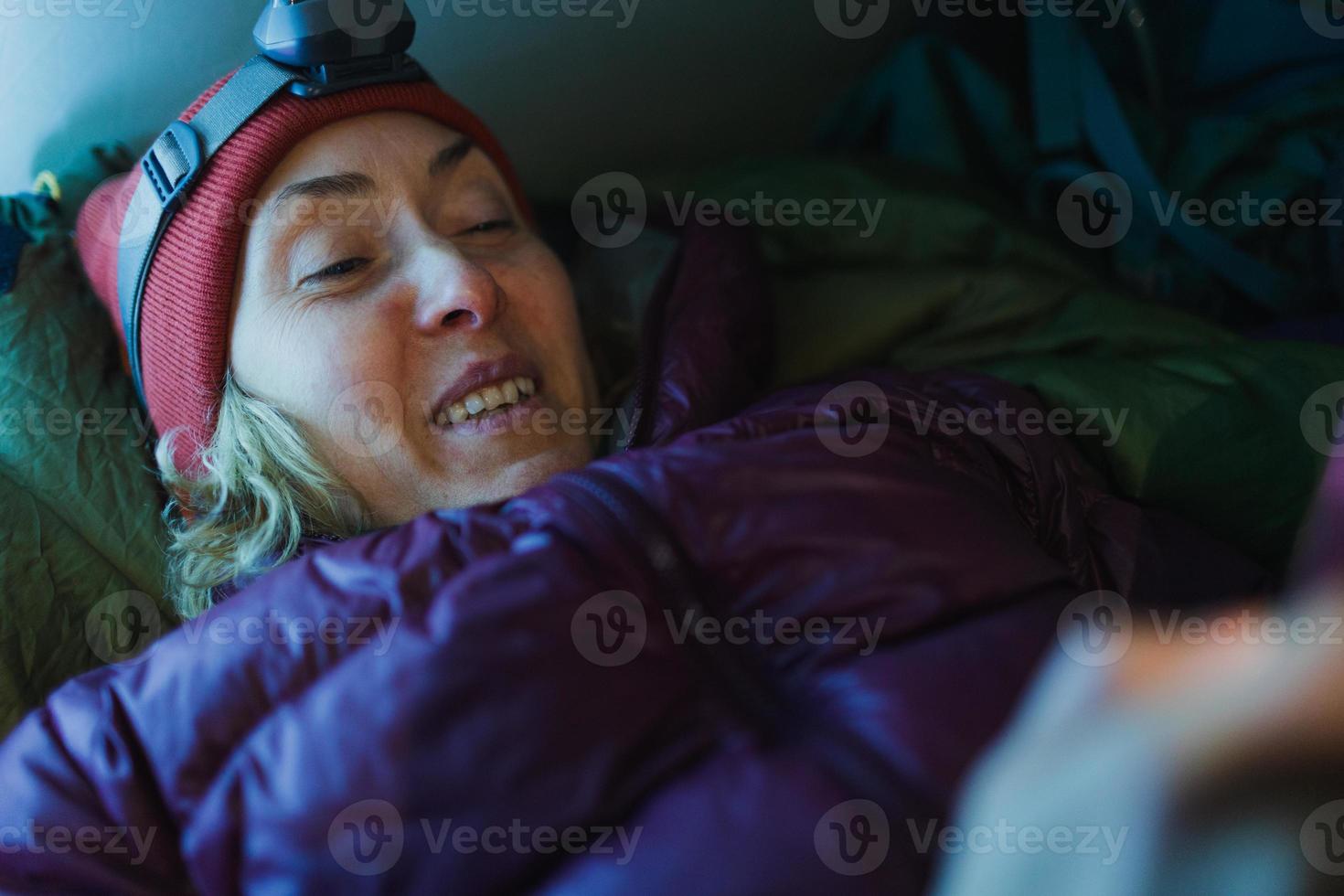uma mulher com um farol está em um saco de dormir em uma barraca e segura um smartphone foto