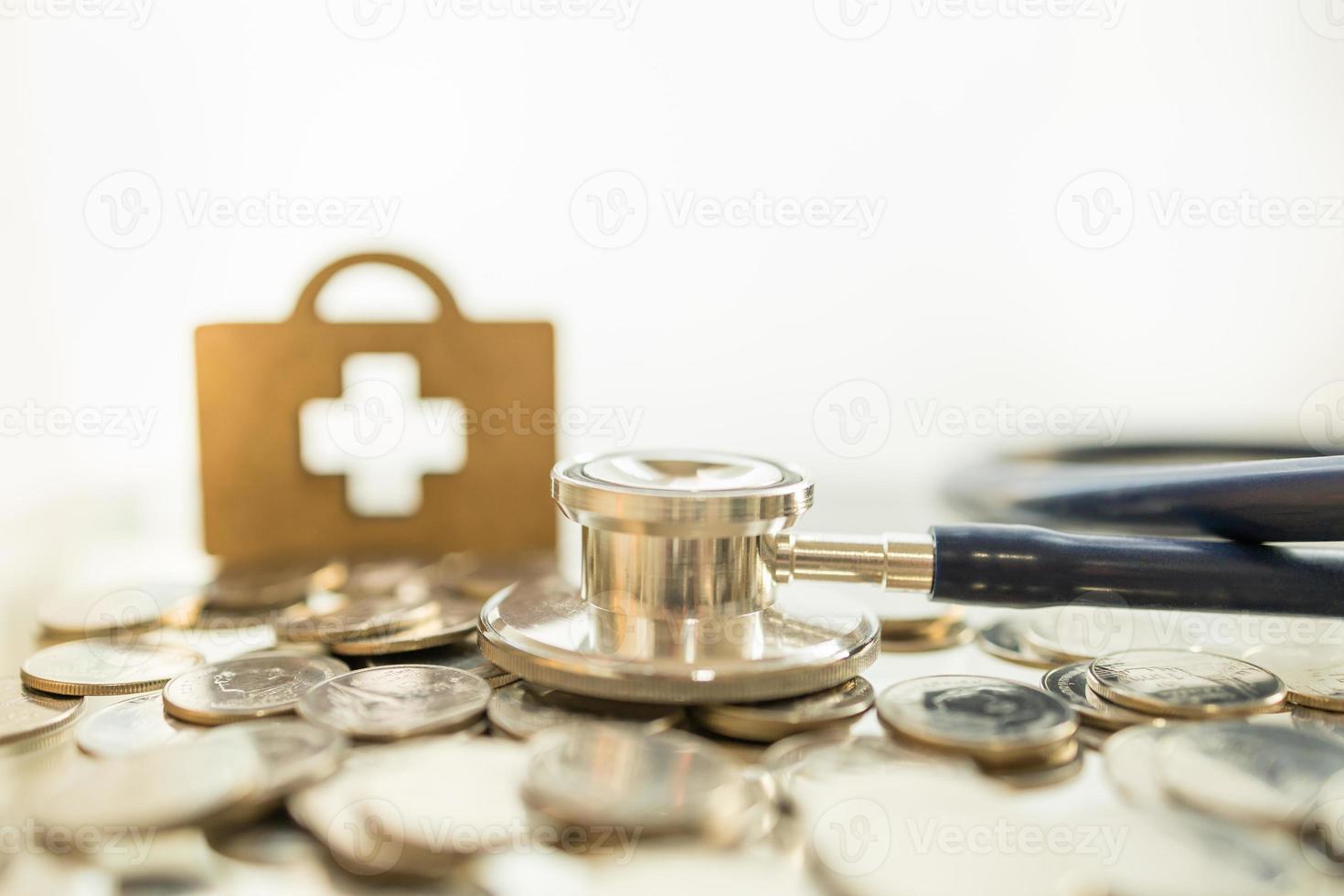 cuidados de saúde e conceito de dinheiro. feche o estetoscópio na pilha de moedas com símbolo de cruz médica de madeira como plano de fundo e espaço de cópia. foto